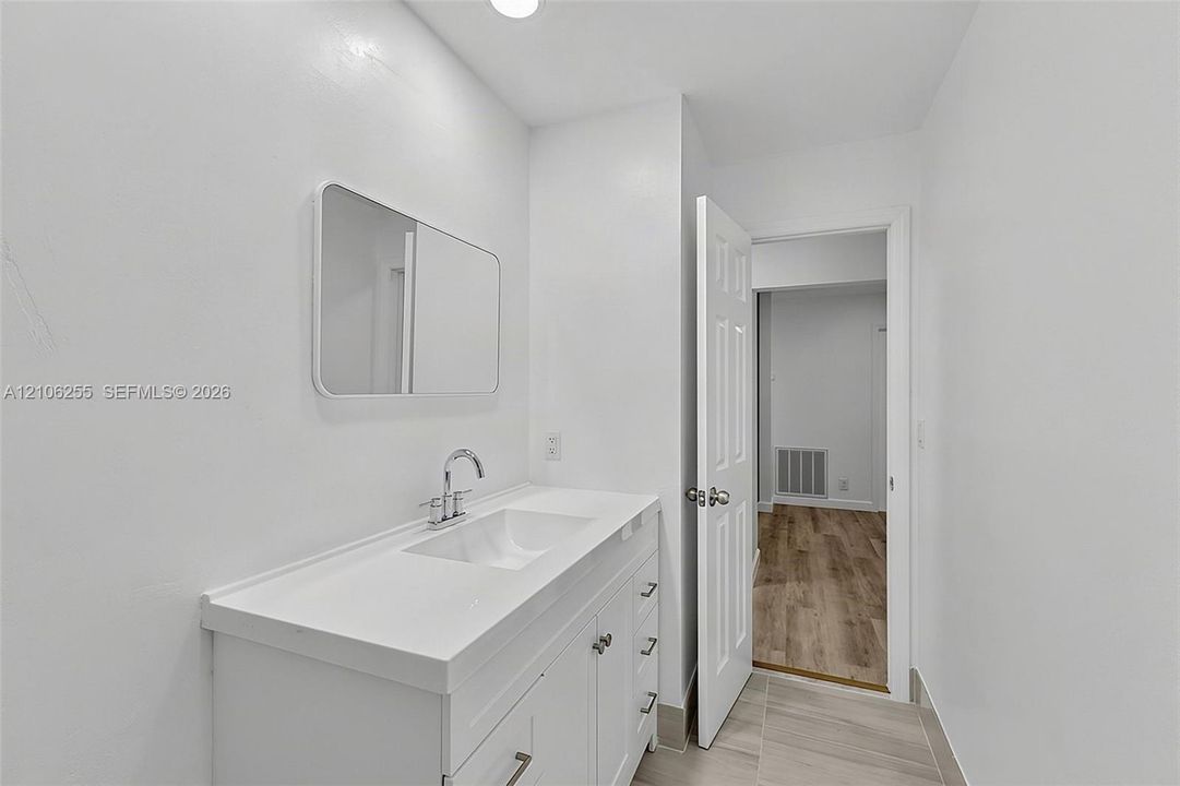 Bathroom vanity with a white composite countertop, integrated rectangular sink, brushed chrome faucet, and rectangular wall-mounted mirror, featuring wood-finish flooring in the surrounding area