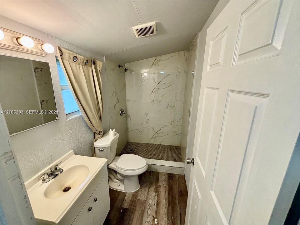 Bathroom featuring wood-finish flooring, a white vanity with an integrated sink, and a walk-in shower with stone-patterned wall tile and a pebble-texture floor