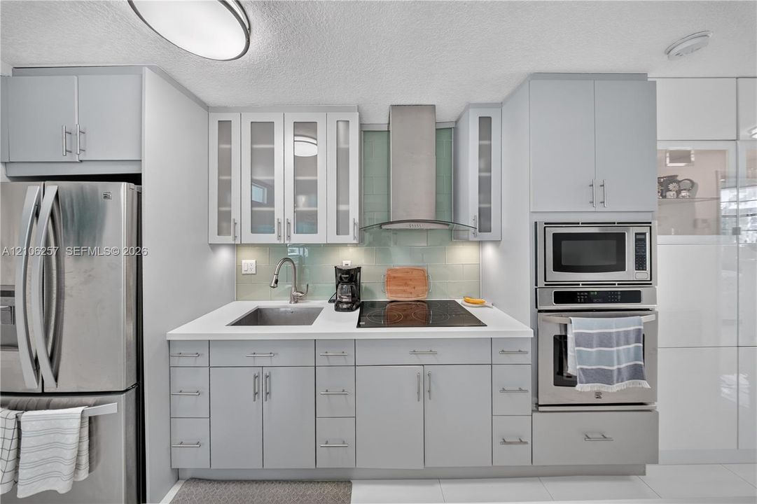 Kitchen featuring light gray cabinetry with glass-front uppers, solid-surface countertops, a glass tile backsplash, and stainless steel built-in appliances including a wall-mounted range hood