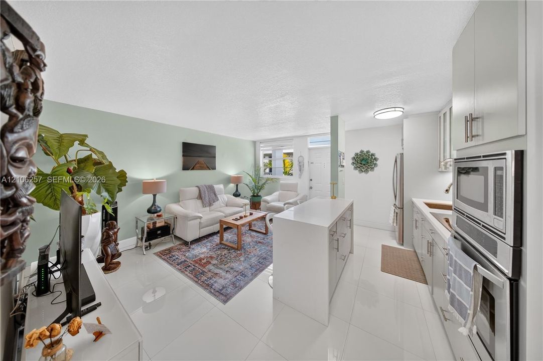 Open-concept living space featuring high-gloss tile flooring, white shaker cabinetry, a central island with a solid-surface countertop, and integrated stainless steel wall oven and microwave