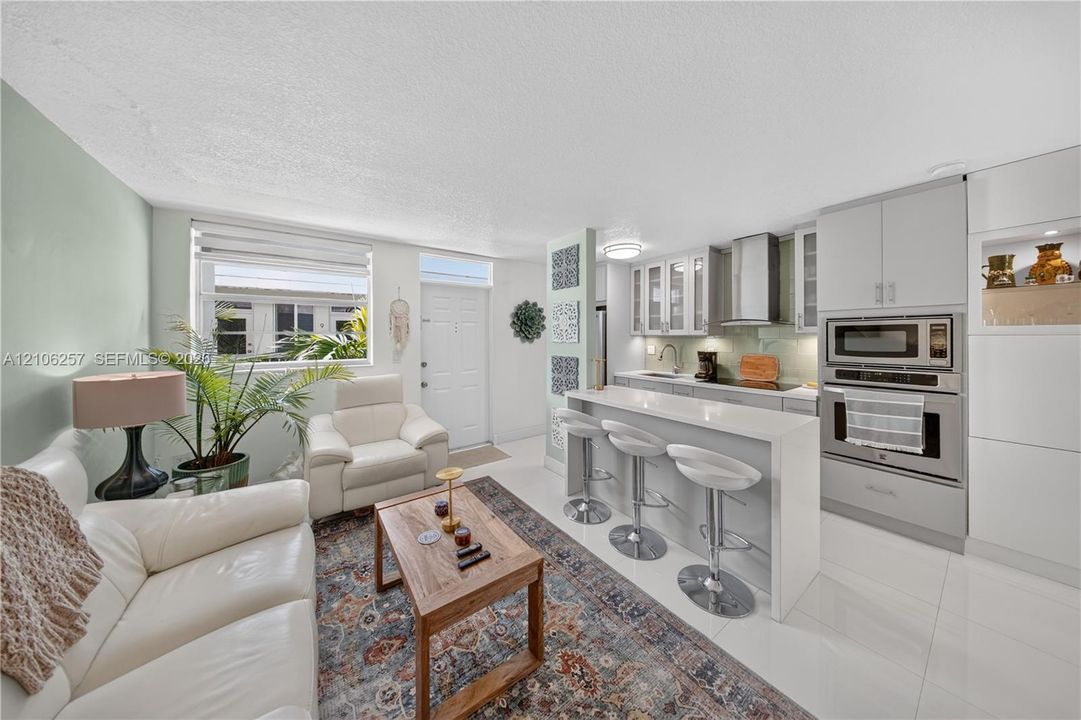 Kitchen featuring light gray cabinetry, a breakfast bar with stone-style countertop, stainless steel built-in wall oven and microwave, stainless steel range hood, and tile flooring