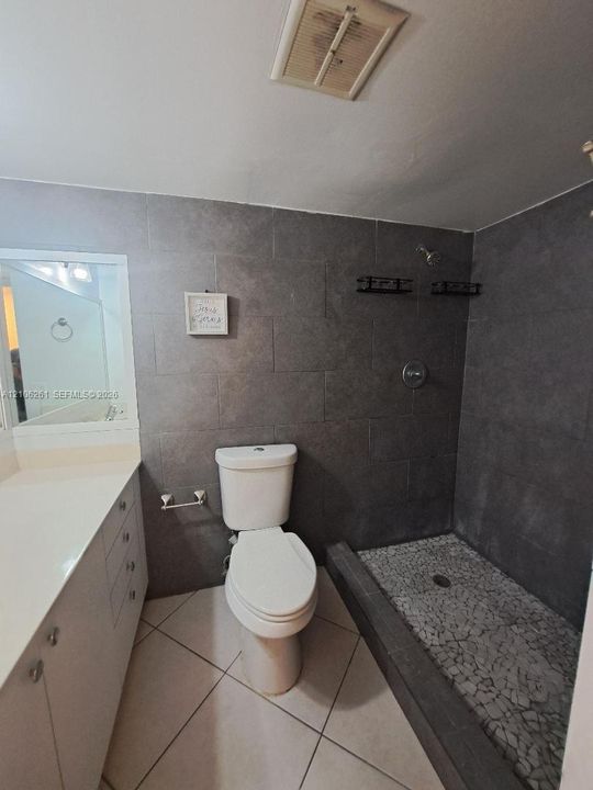 Bathroom featuring large-format dark grey wall tile, a walk-in shower with stone mosaic flooring, a white vanity with a solid-surface countertop, and light-colored diagonal floor tile