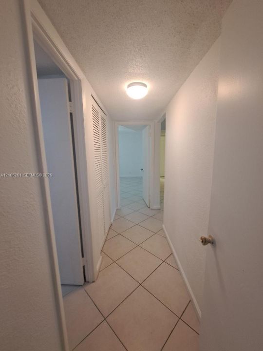 Interior hallway featuring neutral tile flooring laid on a diagonal, crisp white walls, flush-mount ceiling lighting, and louvered-style closet doors