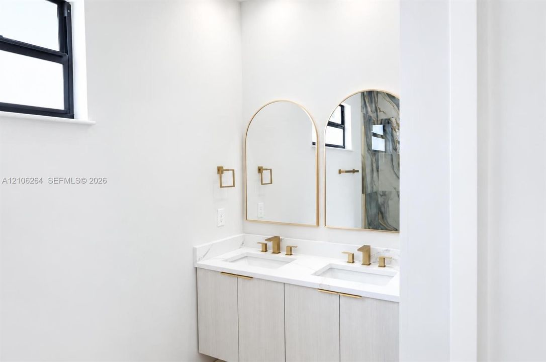 Dual-sink vanity featuring light wood-finish cabinetry, white stone-surfaced countertops, brushed gold-tone faucets, and twin arched mirrors