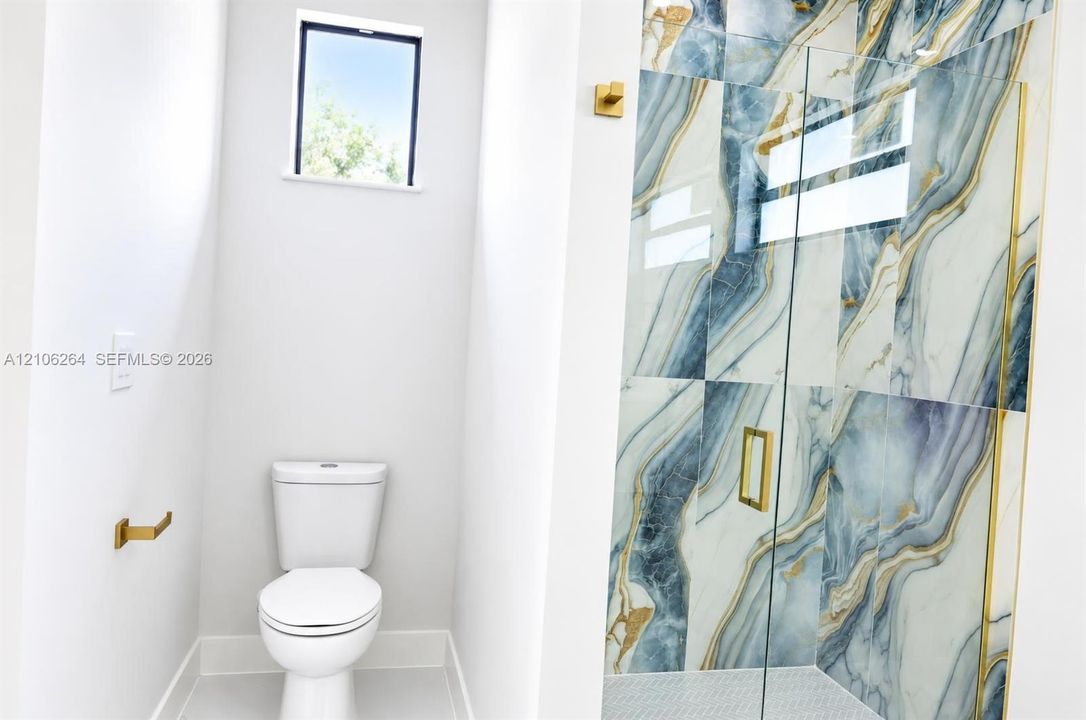 Shower enclosure featuring large-format decorative tile with gold-tone metal trim and hardware, clear glass paneling, and a high-efficiency toilet
