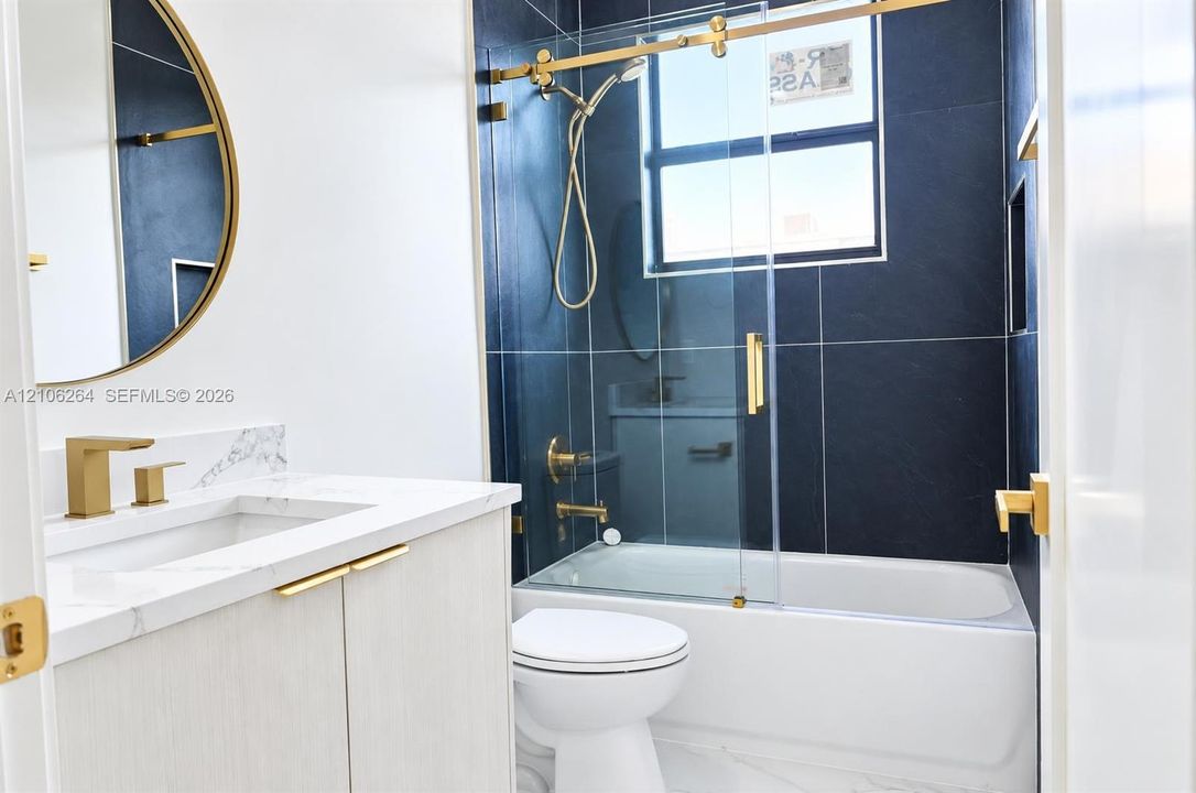 Bathroom featuring a light wood-finish vanity with a stone countertop, a brass-rimmed circular mirror, gold-tone fixtures, and a glass-enclosed shower with dark tile surround