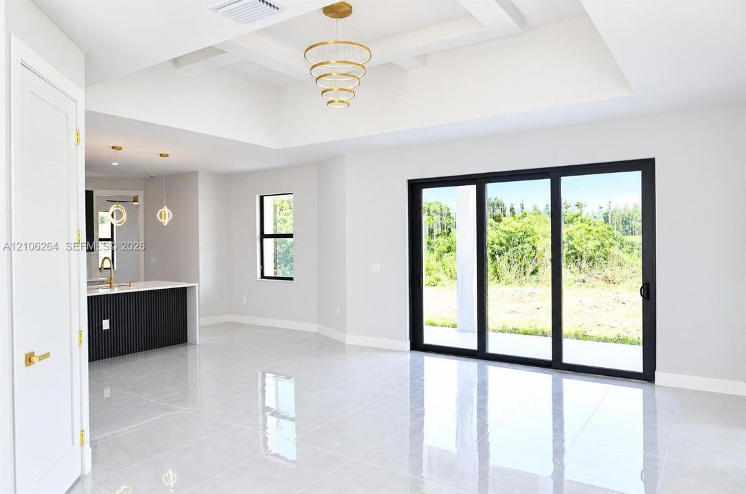 Tray ceiling with multi-tiered gold-tone chandelier, large format polished tile flooring, sliding glass doors with black frames, and kitchen island featuring a black fluted facade