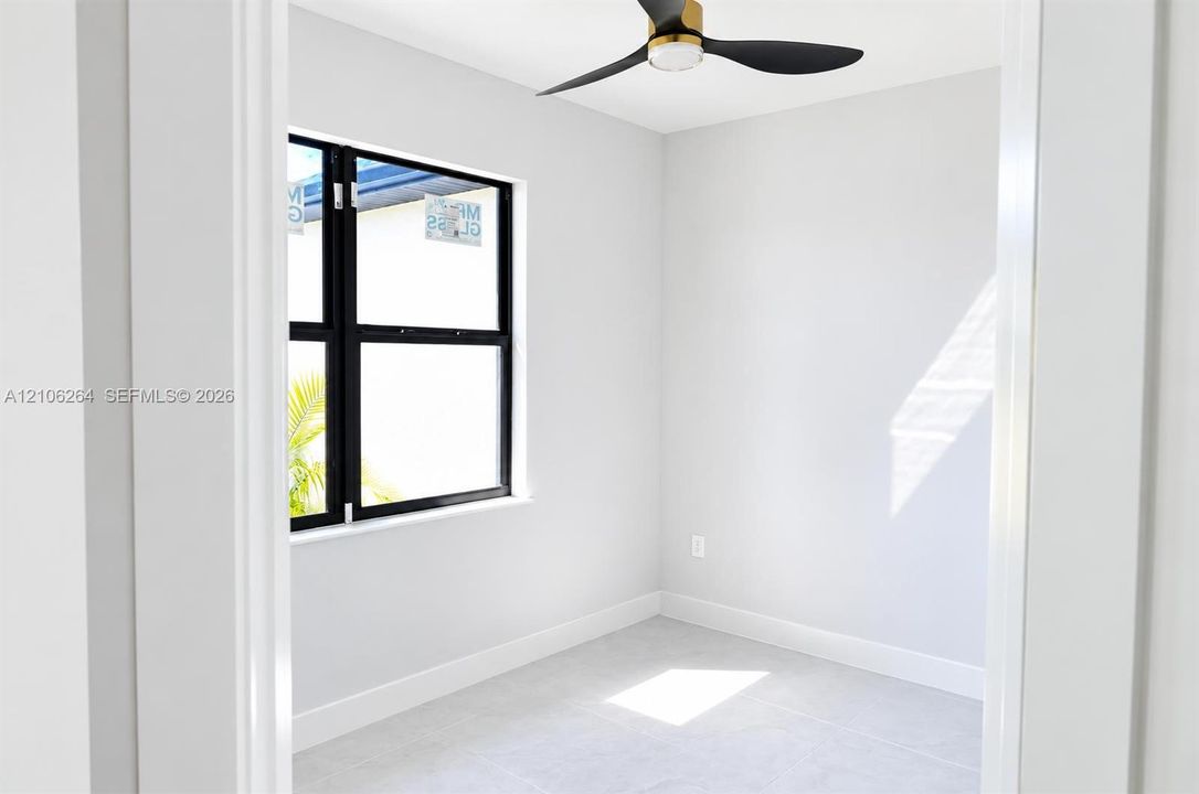 Neutral-toned room featuring a matte black three-blade ceiling fan with gold accents, large-format tile flooring, and a single-hung window with a matte black frame