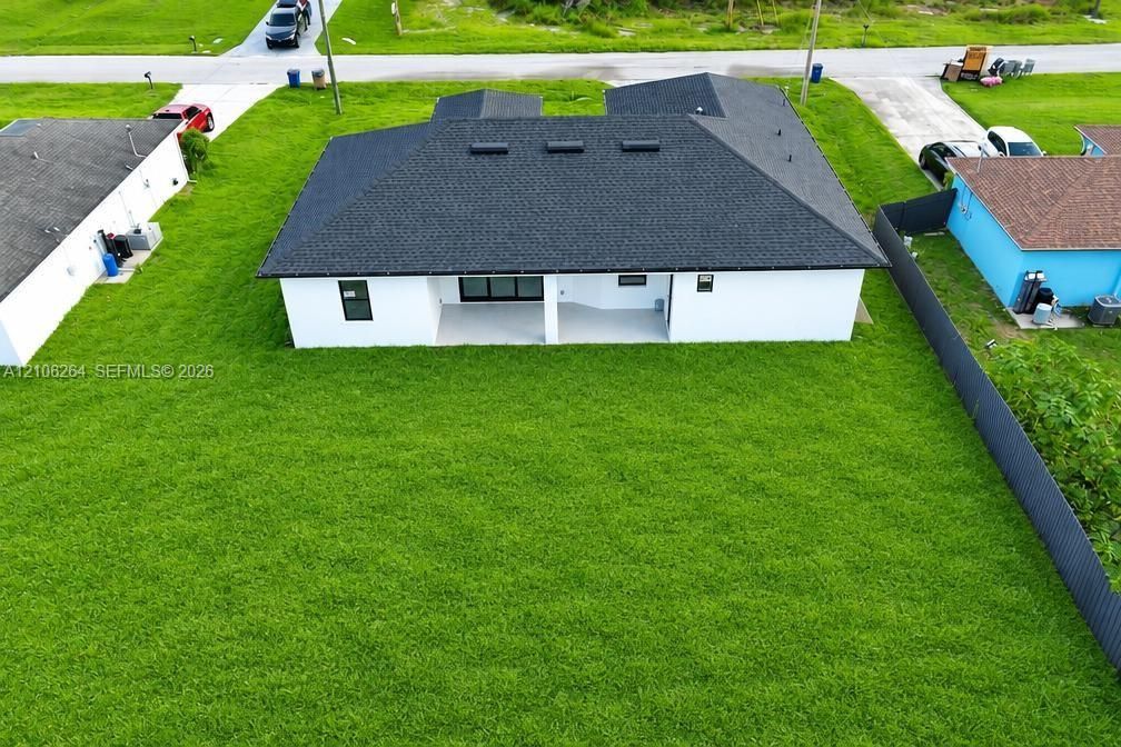 Aerial perspective of a residence featuring a covered patio, expansive lawn, dark shingle roofing, and white exterior wall finish