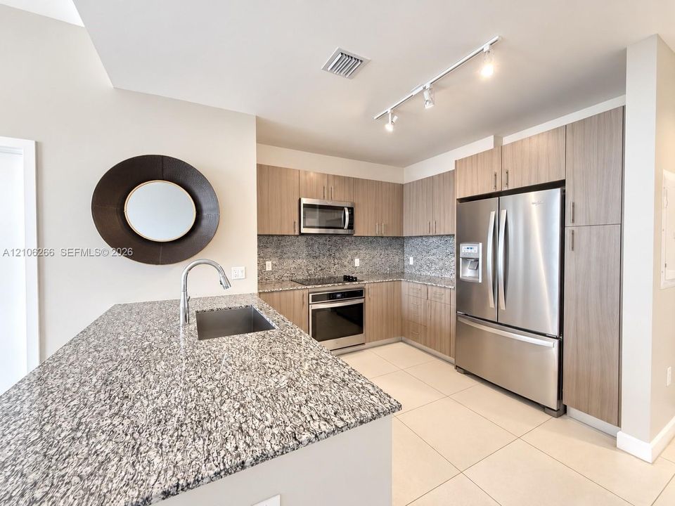Kitchen featuring flat-panel wood-finish cabinetry, speckled stone countertops, stainless steel appliances, a center island with an undermount sink, and tile flooring