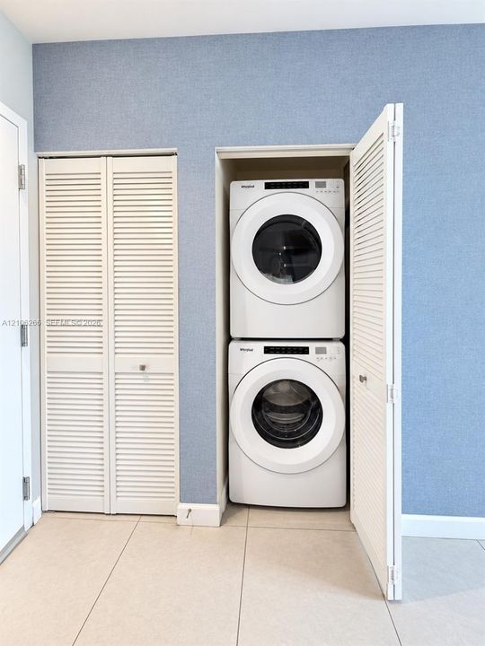 Stacked front-loading laundry center with louvered bi-fold closet doors and neutral tile flooring