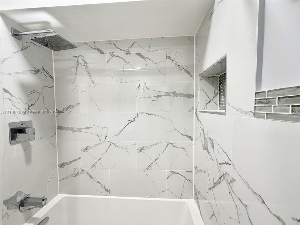 Shower area featuring large-format veined tile surround, recessed shelving niche, glass tile accent border, and chrome rainfall showerhead