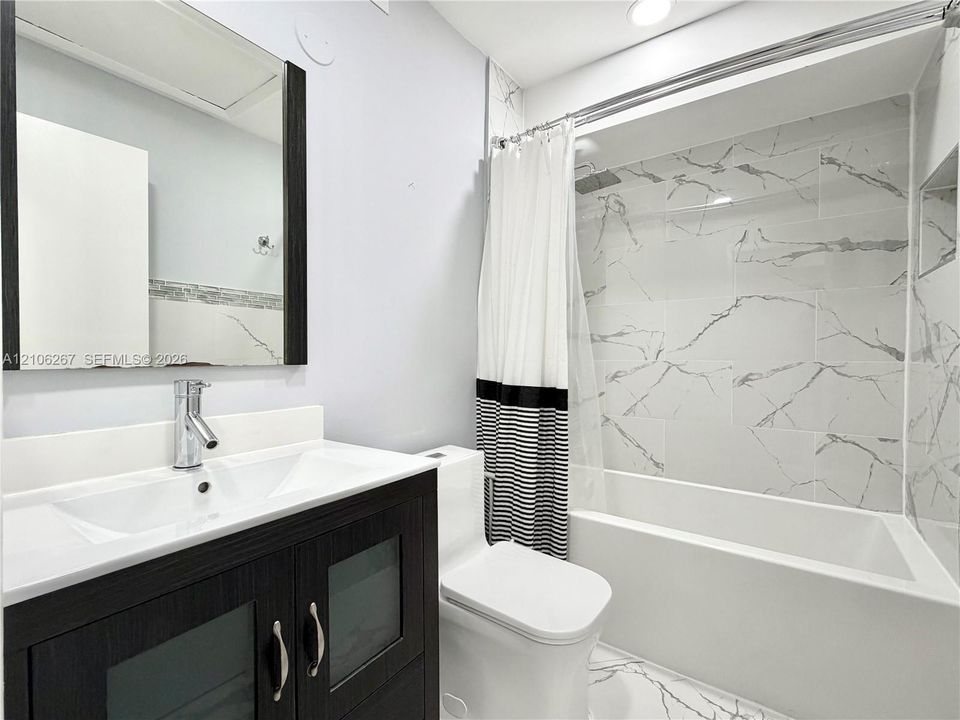 Bathroom featuring a dark wood-finish vanity with frosted glass inserts, a white integrated countertop sink, a full-length framed mirror, and a tub-shower combination with veined tile surround