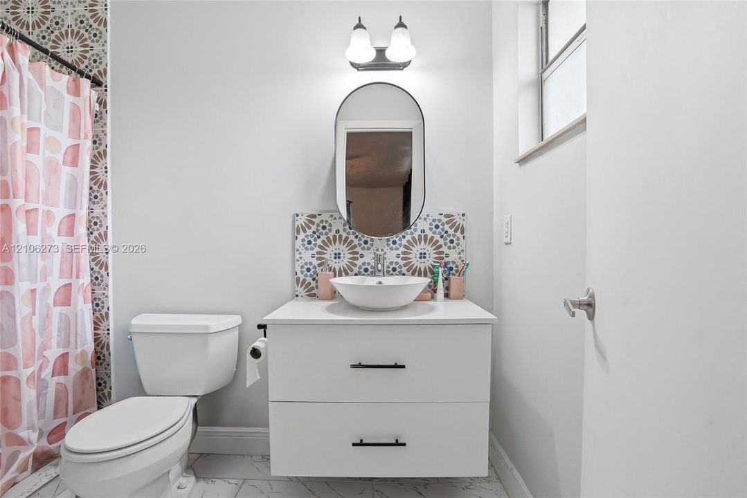 White vanity with a round vessel sink, matte black hardware, oval wall-mounted mirror, decorative geometric tile backsplash, and tile flooring