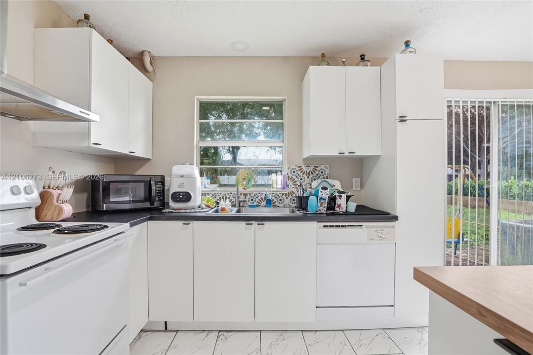 Kitchen featuring white shaker-style cabinetry, matte black countertops, stainless steel sink with a decorative tiled backsplash, white appliance suite, and light-toned tile flooring
