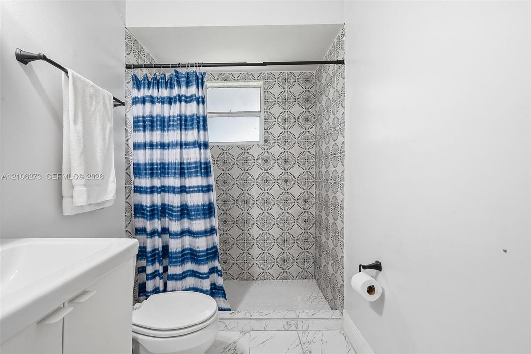 Shower alcove featuring patterned wall tile, white floor tile with gray veining, a built-in horizontal window, a wall-mounted matte black towel bar, and a matte black toilet paper holder