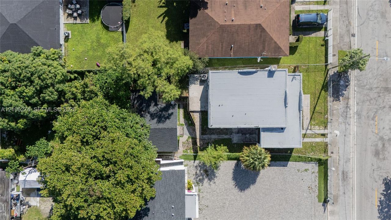 Aerial perspective featuring a flat-roof residence, extensive gravel parking area, mature canopy trees, and fenced perimeter