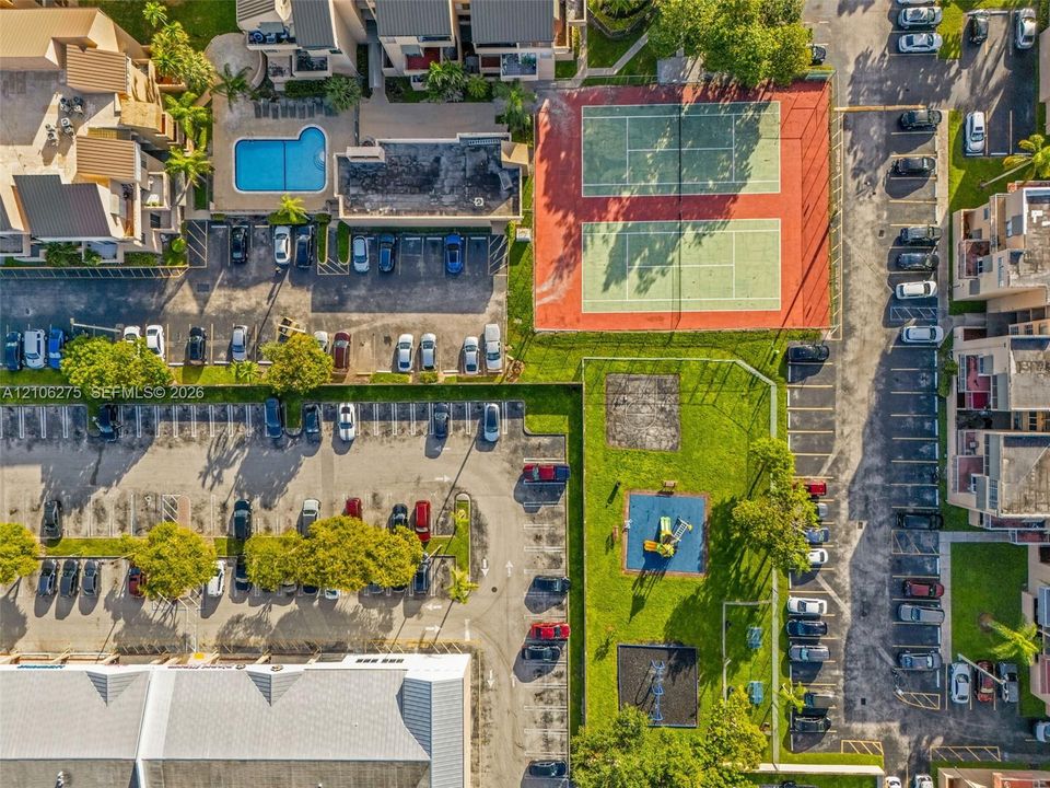 Aerial perspective of community grounds featuring two hard-court tennis surfaces, a swimming pool, a playground area, and designated parking stalls