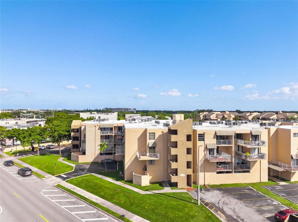 Multi-story residential building featuring a stucco exterior, private balconies, paved surface parking, and manicured surrounding lawns