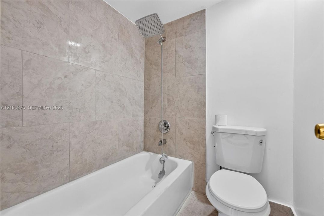 Full-height stone-finish tile surround, square rainfall shower head, chrome wall-mounted fixtures, deep soaking bathtub, and standard-height white toilet