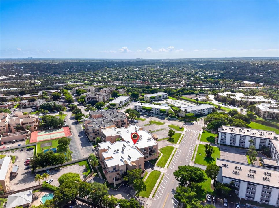 Aerial perspective of a multi-unit complex featuring flat roofing, paved surface parking, tennis courts, and a swimming pool