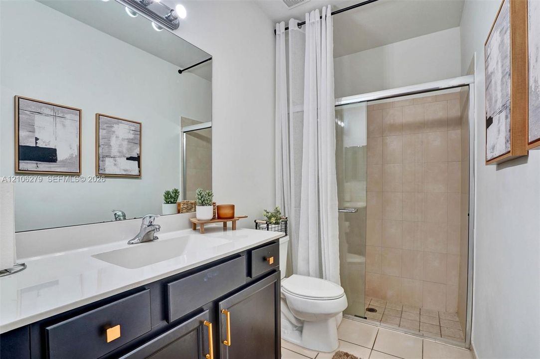 Dark cabinetry vanity featuring gold-toned hardware, white countertop with integrated sink, large wall-mounted mirror, and glass-enclosed shower with tiled surround