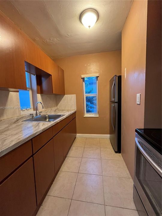 Galley kitchen featuring wood-finish cabinetry, stone-pattern laminate countertops, stainless steel sink with a gooseneck faucet, tile flooring, and stainless steel appliances