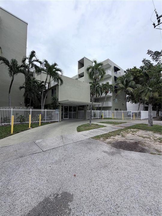 Multi-story building exterior featuring a covered entryway, concrete driveway, white metal fencing, and mature palm trees