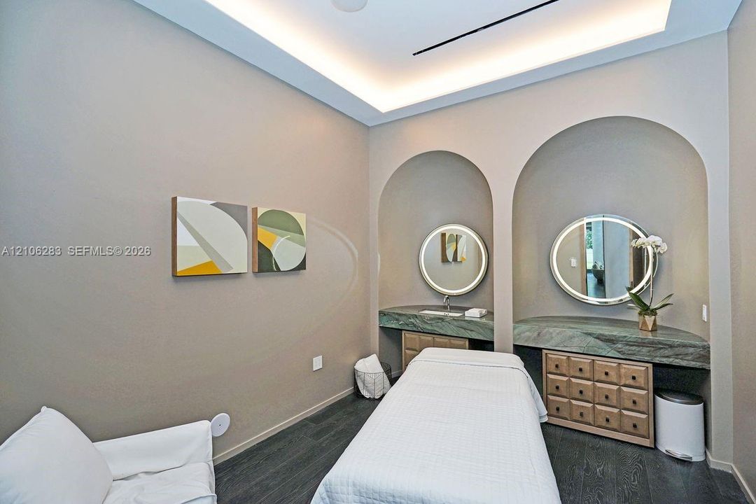 Dual arched wall recesses featuring stone-topped built-in vanities, recessed tray ceiling with integrated perimeter lighting, round back-lit mirrors, and dark-toned plank flooring
