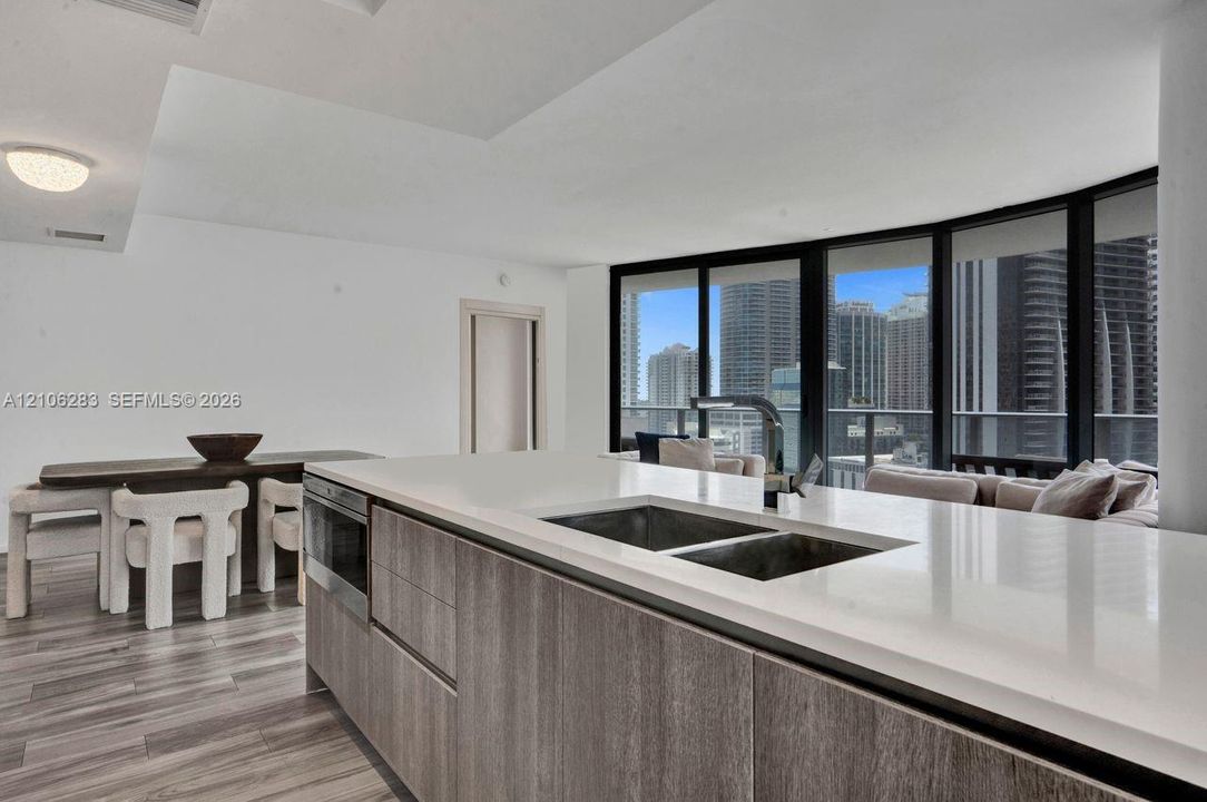 Kitchen island featuring wood-finish cabinetry, a solid surface countertop, an undermount double-basin sink, and a gooseneck faucet, set against floor-to-ceiling windows with urban scenery