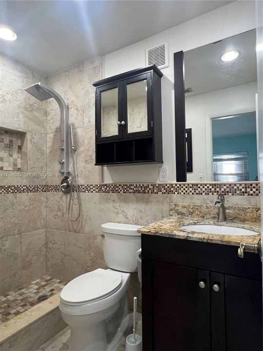 Full-tile shower stall with integrated recessed shelving, multi-head shower system, stone-finish vanity countertop with undermount sink, dark-finish cabinetry, and framed mirror