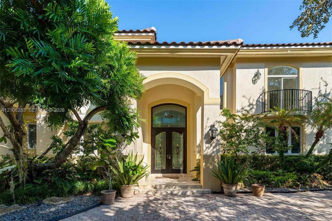 Stucco exterior featuring an arched entryway with decorative glass double doors, a second-story balcony with metal railing, and a stone-paver driveway