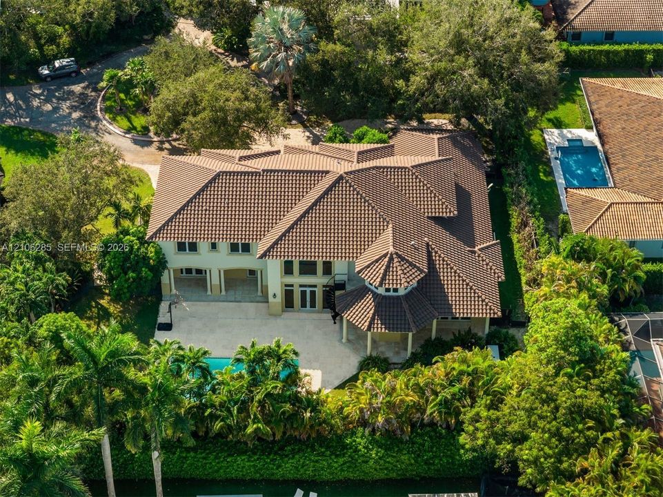Aerial view of a two-story residence featuring a tile roof, light-toned exterior finish, stone-paved patio, private swimming pool, and dense perimeter landscaping