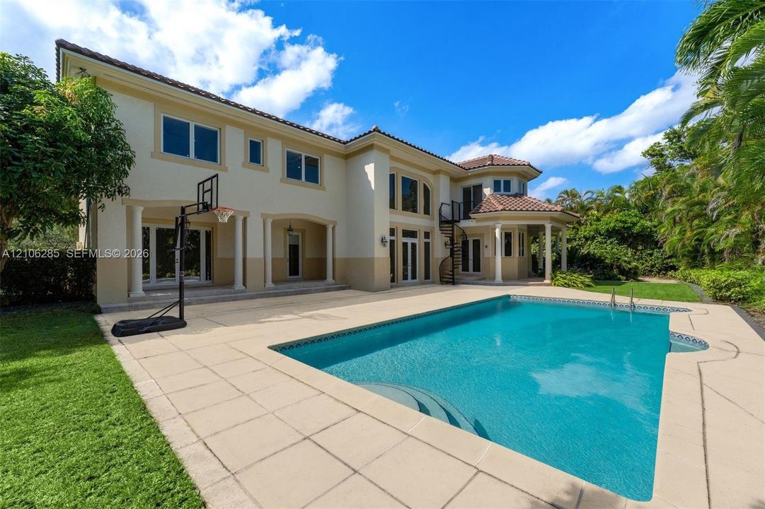 Two-story residence featuring a tile roof, expansive stone-paved pool deck, custom swimming pool, covered patio with structural columns, and a spiral staircase
