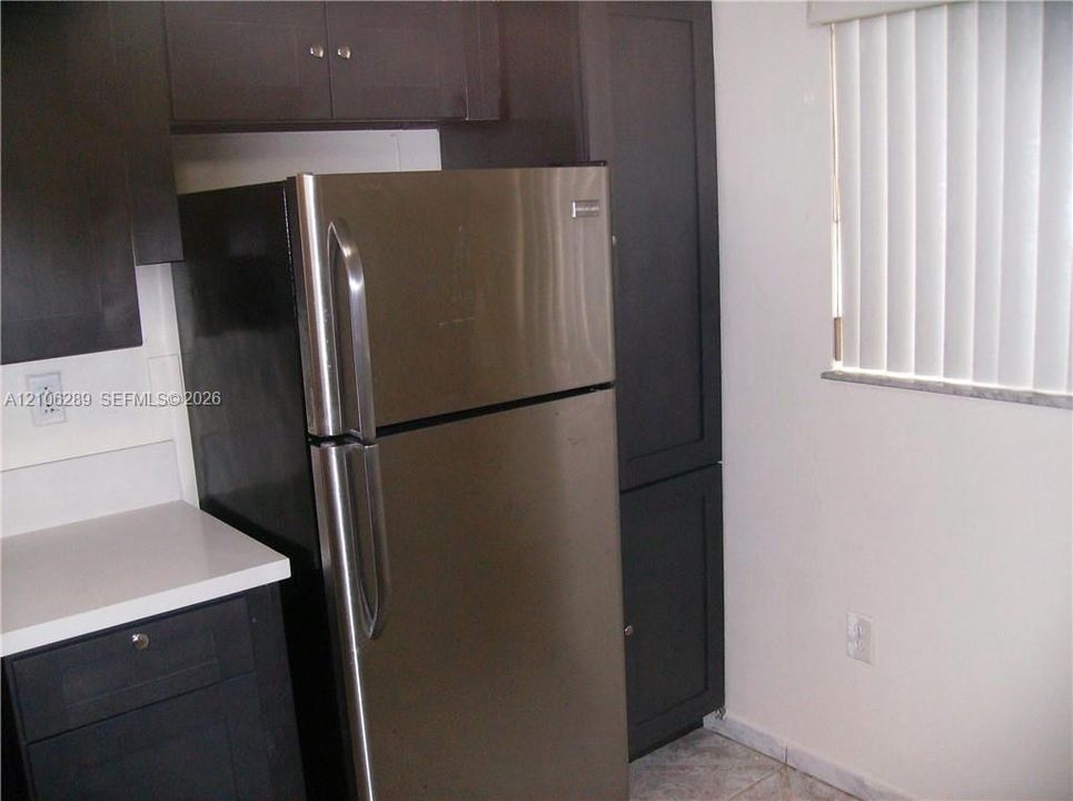 Dark-toned cabinetry, stainless steel refrigerator, white solid-surface countertops, and vertical window blinds