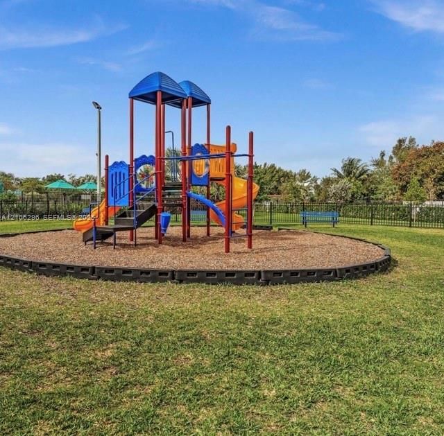 Outdoor recreational area featuring a multi-level playground structure, wood chip surface, metal perimeter fencing, and open green space