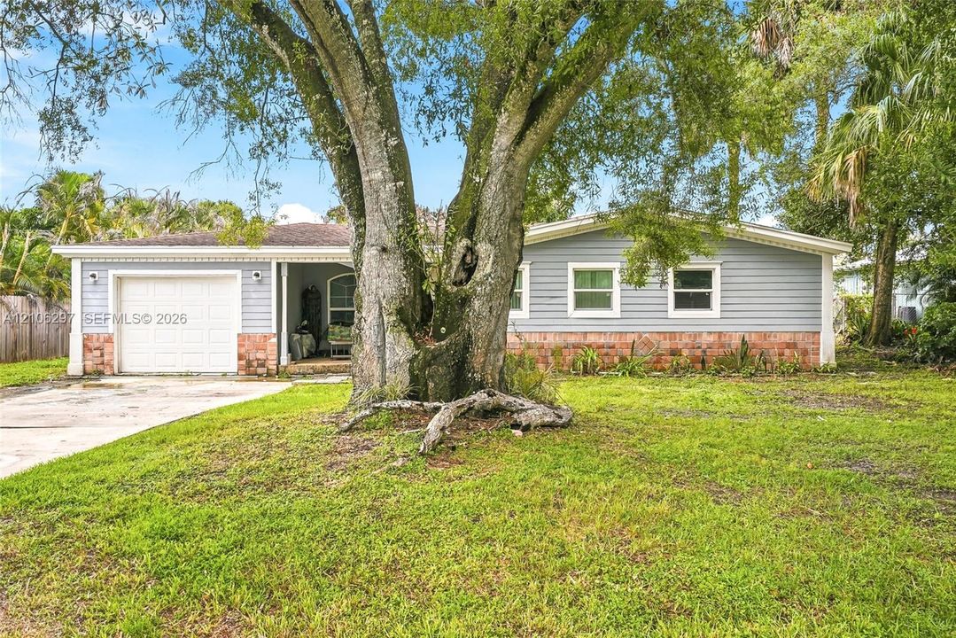 Single-story residence featuring light blue horizontal siding, red brick masonry accents, an attached garage, and a mature tree on the front lawn