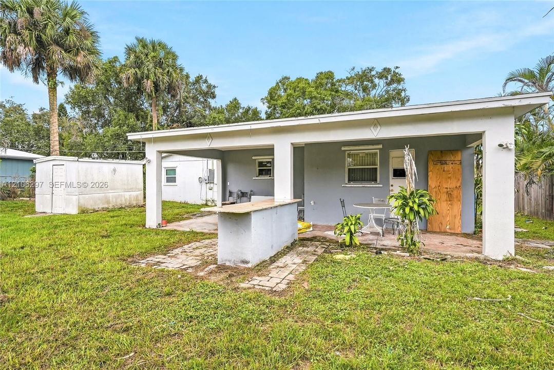 Covered concrete patio featuring an integrated masonry bar, standalone storage shed, and mature palm trees
