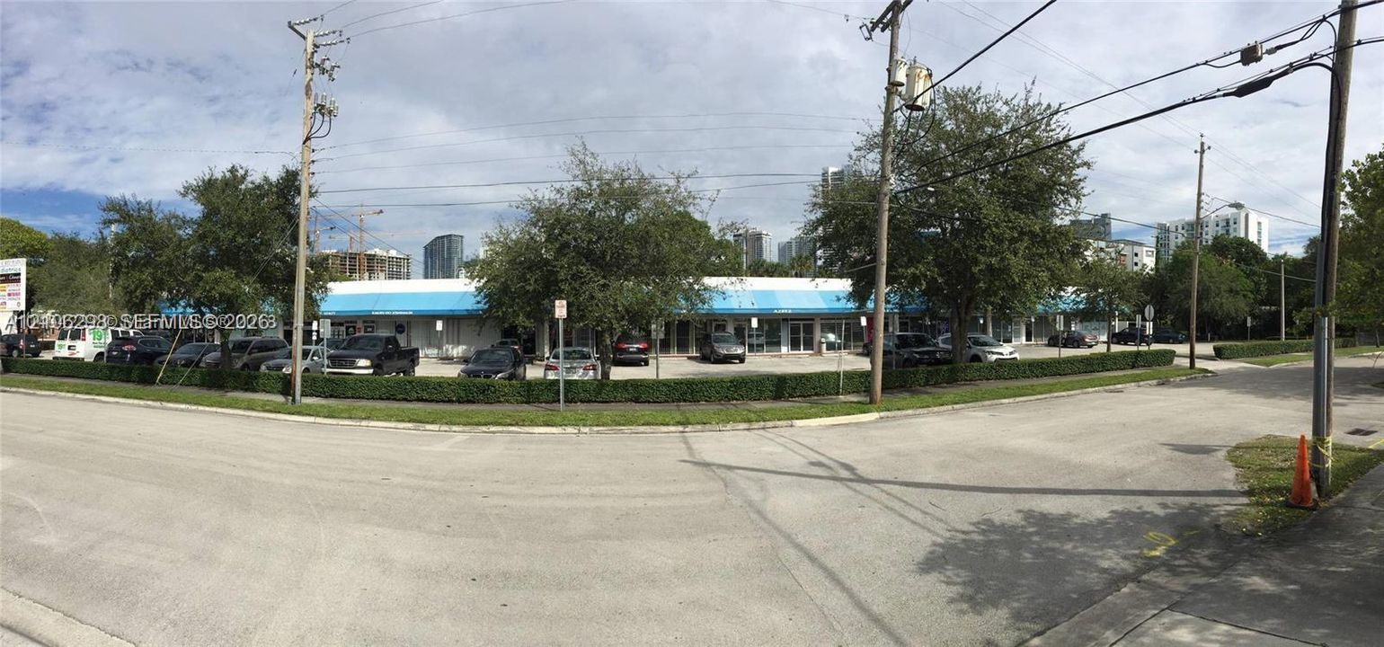 Single-story commercial plaza featuring an expansive surface parking lot, blue metal awnings, glass storefronts, and manicured perimeter hedging