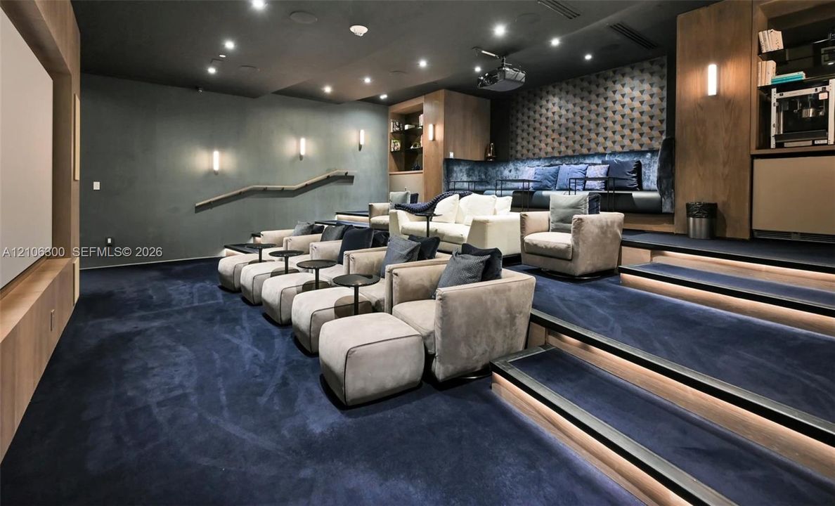 Media room featuring tiered flooring, recessed stair lighting, integrated wooden cabinetry, a projector, and wall-mounted sconce lighting