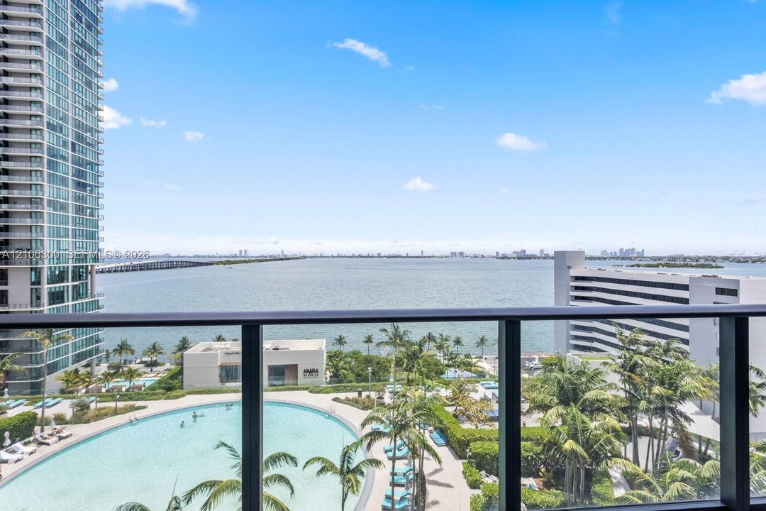 Balcony featuring a dark metal railing, overlooking a large swimming pool, landscaped grounds with mature palm trees, and coastal water