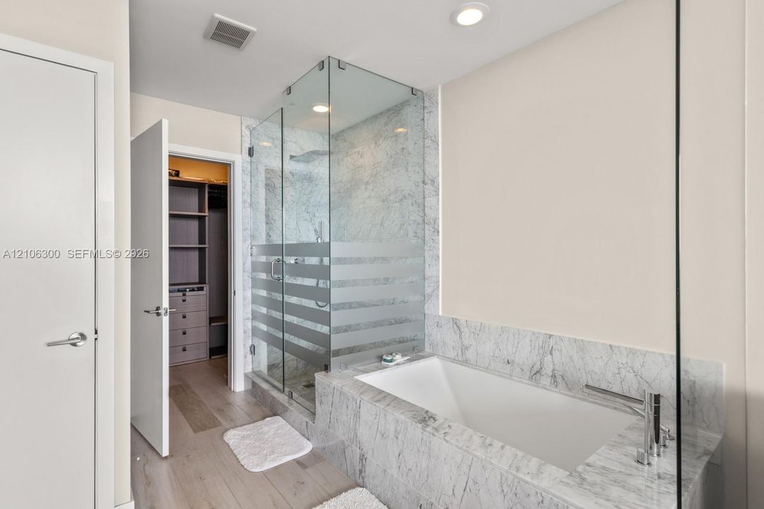 Stone-surround soaking tub with deck-mounted faucet, glass-enclosed shower, wood-finish flooring, and walk-in closet with built-in cabinetry
