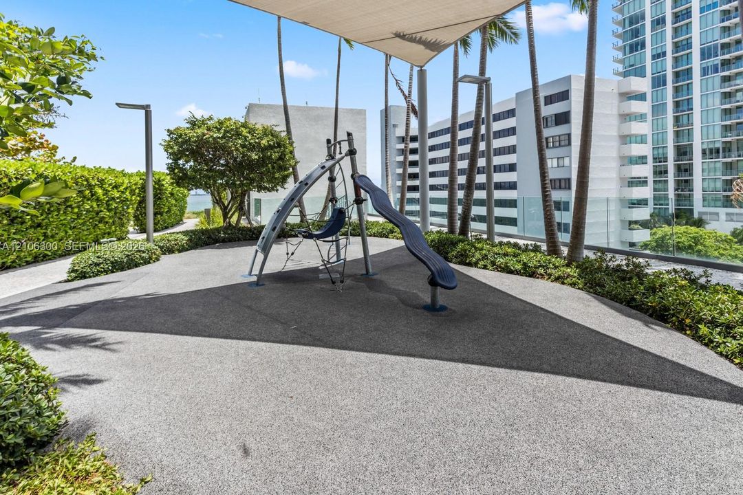 Outdoor recreation area featuring a play structure with a slide, rubberized surfacing, overhead shade canopy, perimeter landscaping, and vertical lighting fixtures