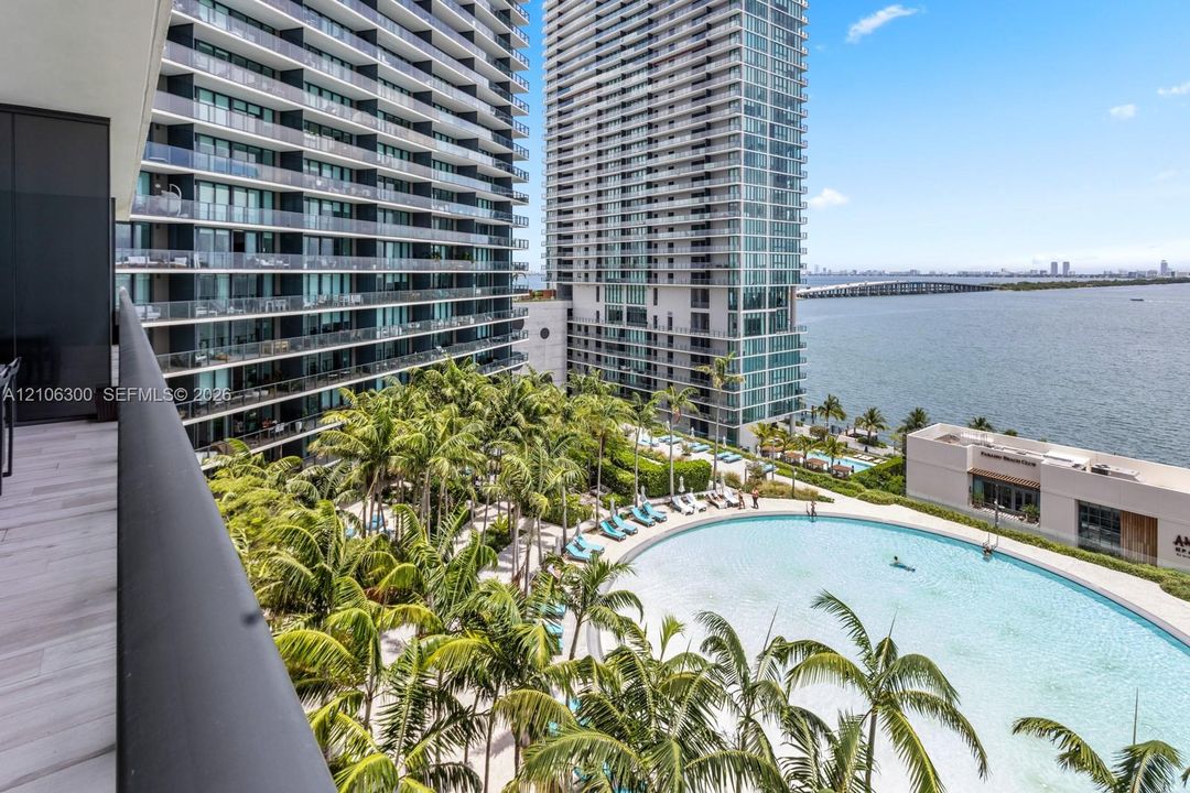 Private balcony with wood-finish flooring, overlooking a large swimming pool, landscaped palm trees, and waterfront horizon