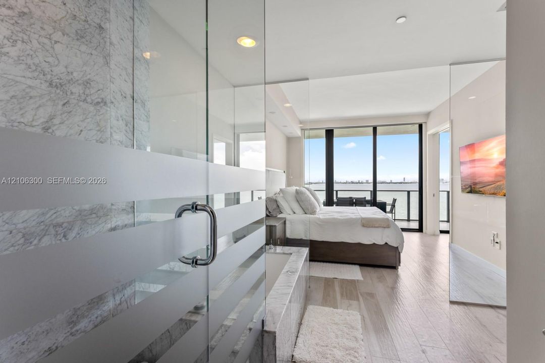 Floor-to-ceiling windows, stone-clad soaking tub, glass-enclosed shower with frosted detailing, wood-finish flooring, and recessed lighting