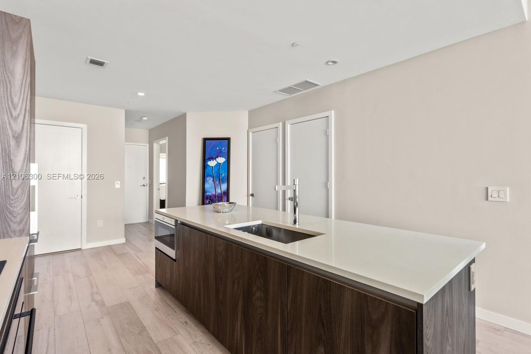Kitchen island featuring dark wood-finish cabinetry, a white countertop, an integrated stainless steel undermount sink, and a high-arc faucet, complemented by light wood-finish flooring