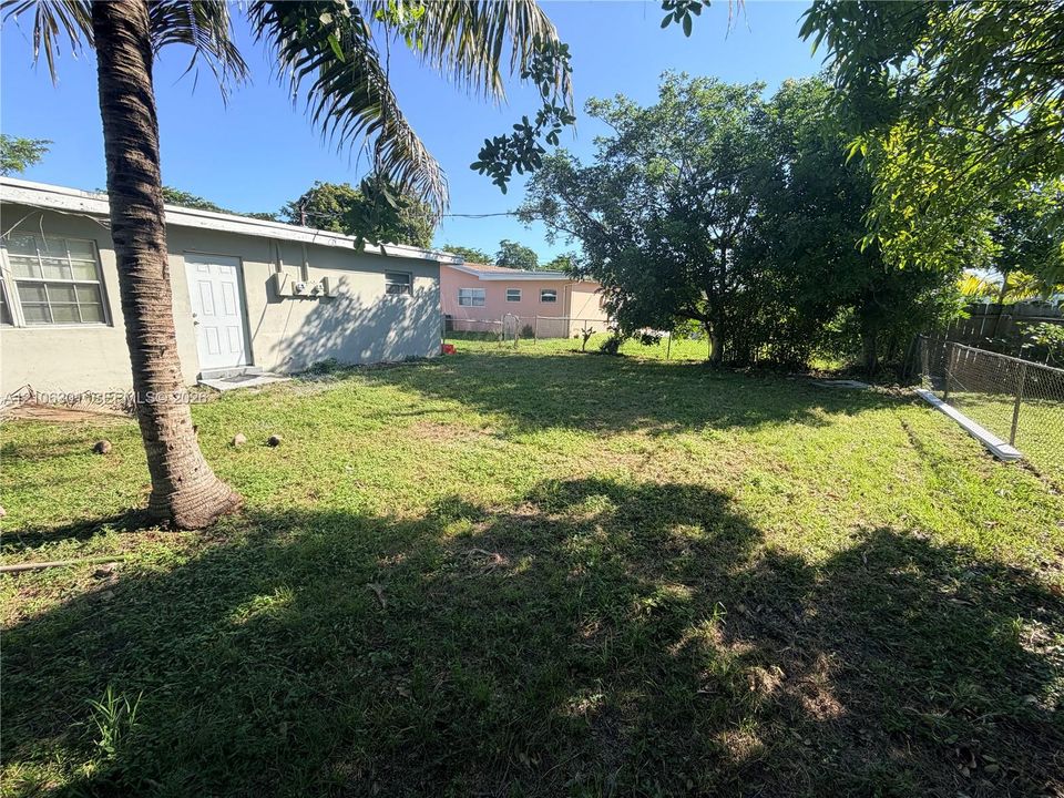 Rear exterior featuring a grass lawn, mature trees, chain-link fencing, and a palm tree