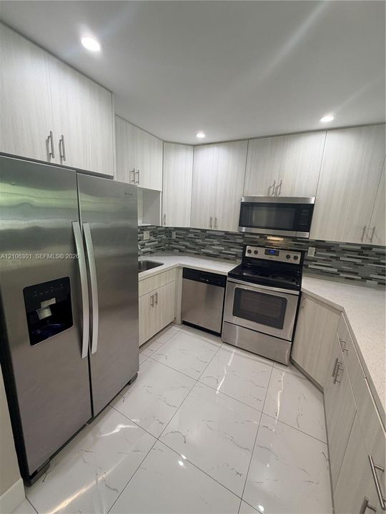 Kitchen featuring light-toned cabinetry, stainless steel appliances, stone-finish countertops, decorative tile backsplash, and large-format tile flooring