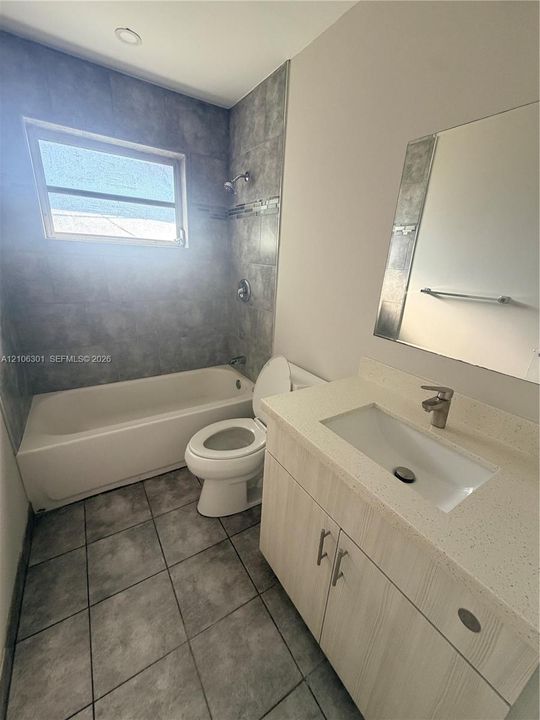 Bathroom featuring a light wood-finish vanity with a solid-surface countertop, rectangular undermount sink, tiled surround shower-tub combination, and floor tiling