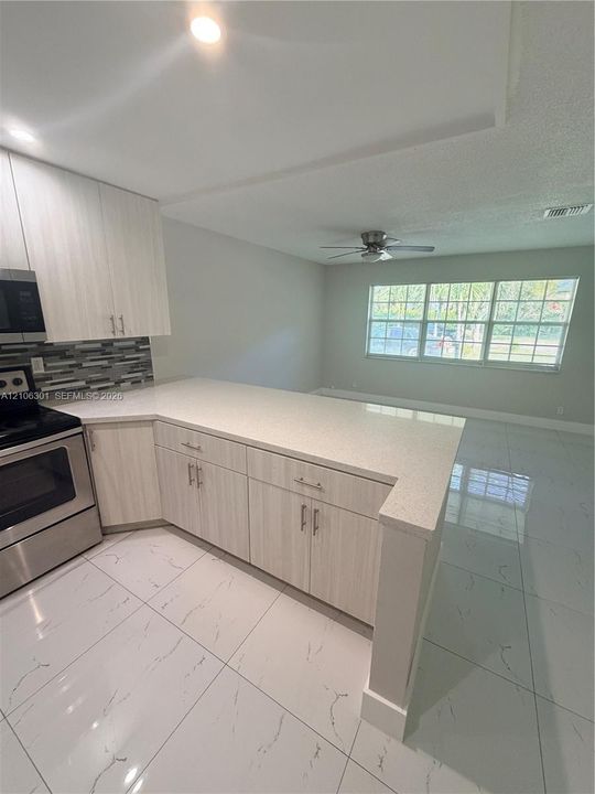 Kitchen featuring light-toned cabinetry, solid-surface countertops, decorative mosaic tile backsplash, stainless steel appliances, and large-format porcelain tile flooring